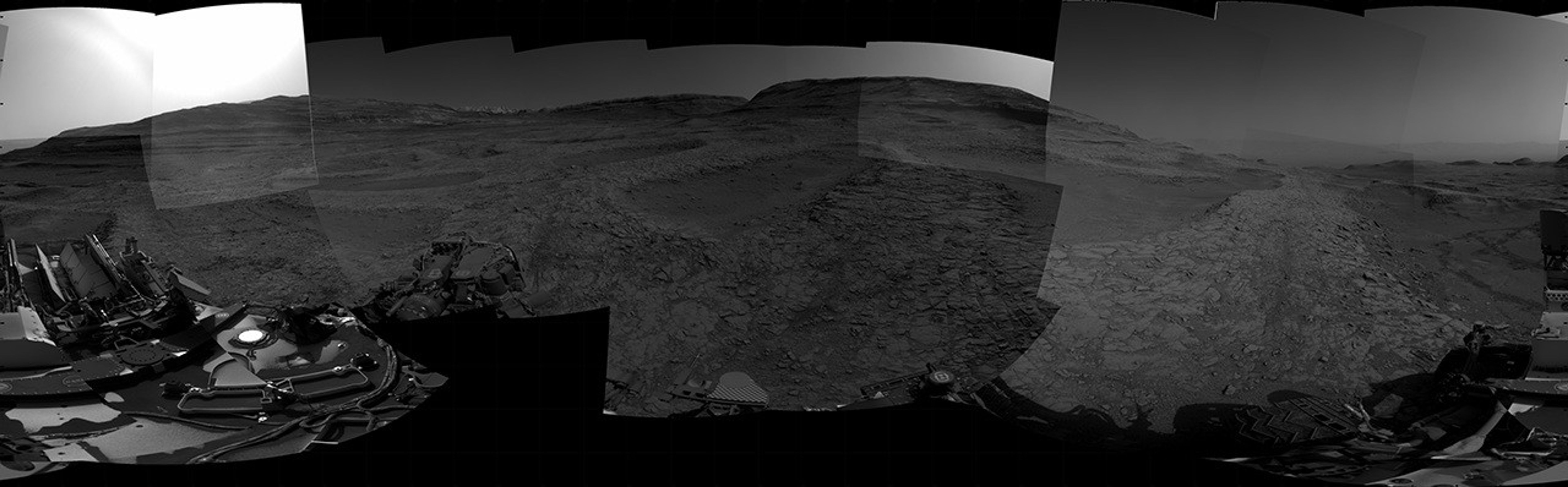 A grayscale panorama of the Martian surface, possibly at night or in twilight, shows a wide field of flat, dark gray terrain dotted with multiple jagged, medium-sized rocks stretching into the distance, where hills rise from the ground on the horizon. At center-left, a bright light behind a hill illuminates part of the sky, while the rest of the scene is fairly dark. Portions of the Curiosity rover are visible along the bottom of the image, including wheels in the lower-right corner.