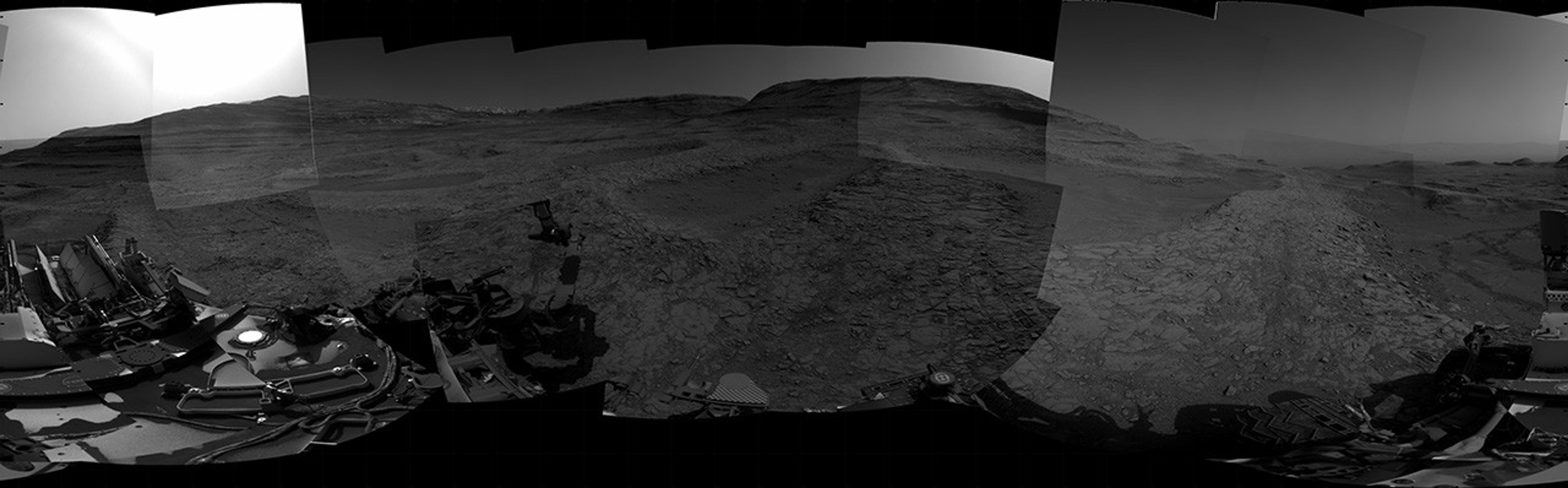 A grayscale panorama of the Martian surface, possibly at night or in twilight, shows a wide field of flat, dark gray terrain dotted with multiple jagged, medium-sized rocks stretching into the distance, where hills rise from the ground on the horizon. At center-right, a bright light behind a hill illuminates part of the sky, while the rest of the scene is fairly dark. Portions of the Curiosity rover are visible along the bottom of the image, including wheels in the lower-right corner.