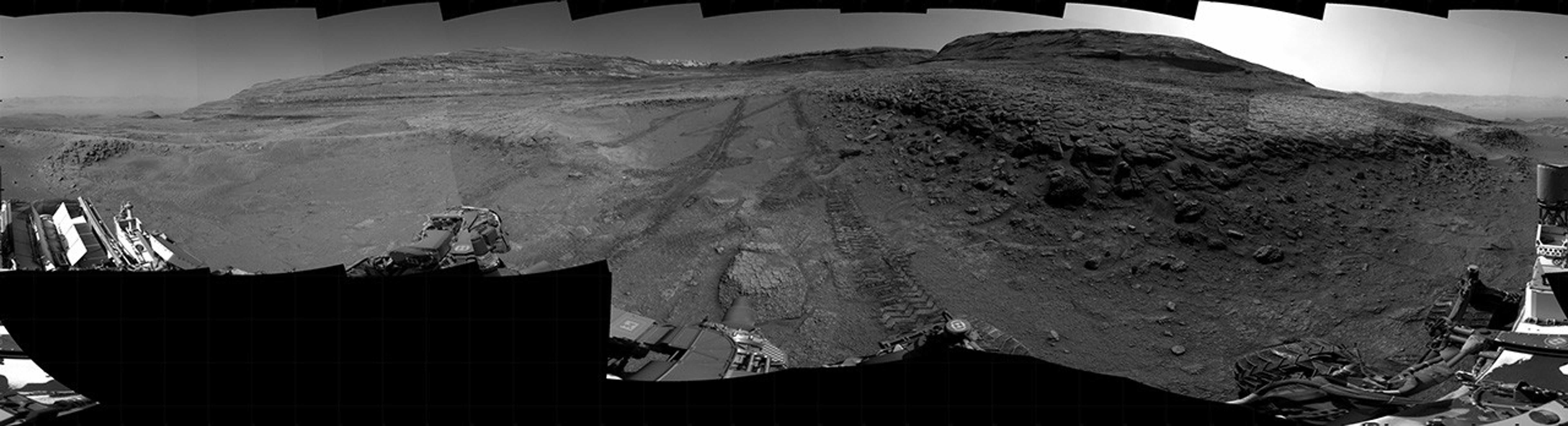 A grayscale panorama of the Martian surface, possibly at night or in twilight, shows a wide field of flat, dark gray terrain dotted with multiple jagged, medium-sized rocks stretching into the distance, where hills rise from the ground on the horizon. At center-right, a bright light behind a hill illuminates part of the sky, while the rest of the scene is fairly dark. Portions of the Curiosity rover are visible along the bottom of the image, including wheels in the lower-right corner.