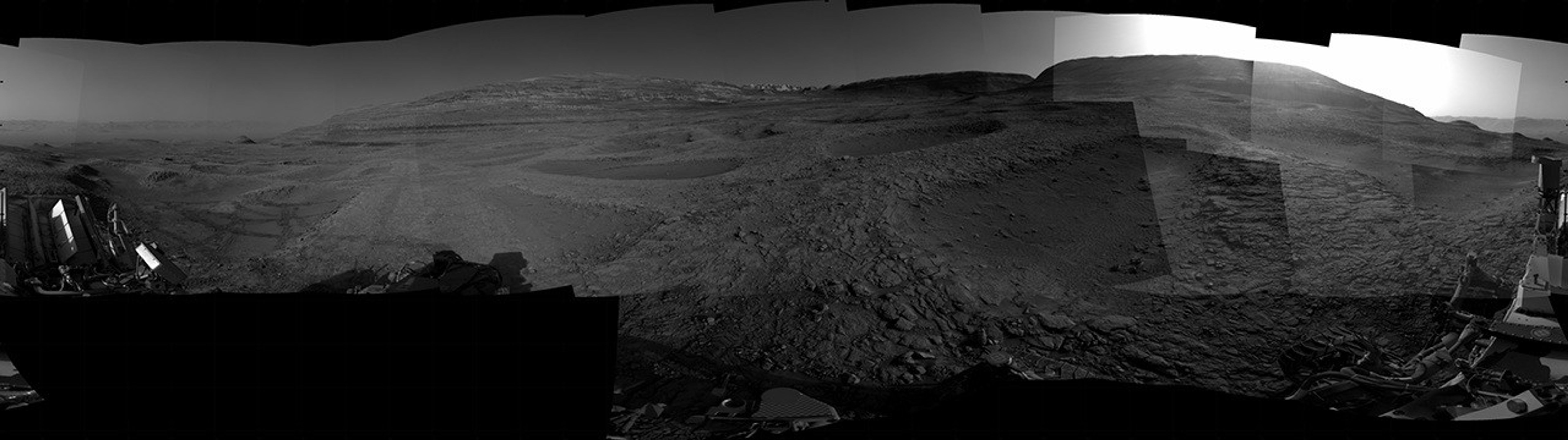 A grayscale panorama of the Martian surface, possibly at night or in twilight, shows a wide field of flat, dark gray terrain dotted with multiple jagged, medium-sized rocks stretching into the distance, where hills rise from the ground on the horizon. At center-right, a bright light behind a hill illuminates part of the sky, while the rest of the scene is fairly dark. Portions of the Curiosity rover are visible along the bottom of the image, including wheels in the lower-right corner.