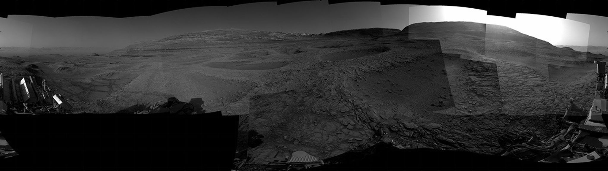 A grayscale panorama of the Martian surface, possibly at night or in twilight, shows a wide field of flat, dark gray terrain dotted with multiple jagged, medium-sized rocks stretching into the distance, where hills rise from the ground on the horizon. At center-right, a bright light behind a hill illuminates part of the sky, while the rest of the scene is fairly dark. Portions of the Curiosity rover are visible along the bottom of the image, including wheels in the lower-right corner.