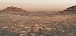 Rover track marks cut through dusty, rocky reddish-brown terrain on Mars, flanked by rounded hills and distant mountains under a hazy sky.
