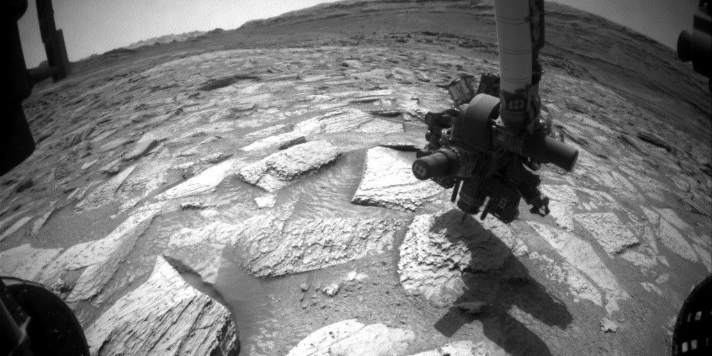 A black-and-white, wide-angle photograph taken on Mars by the Curiosity rover. In the foreground, the rover's robotic arm with a complex cluster of instruments at its end reaches down, casting a dark shadow onto the ground. The terrain consists of flat, jagged, light-colored rock slabs separated by patches of dark sand with wind-blown ripples. Parts of the rover's hardware are visible in the bottom corners of the frame. In the background, a desolate, rocky landscape stretches toward a curved horizon lined with distant hills under a pale sky.