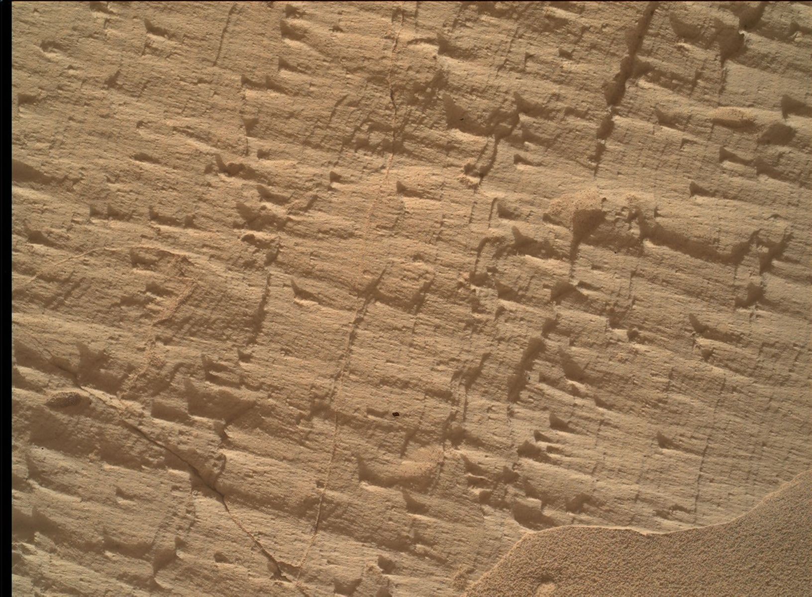 A close-up color photograph taken by the Curiosity rover showing a rough, tan-colored rock surface on Mars. The rock is highly textured, covered in fine parallel layers, small raised ridges, and thin cracks that cast tiny shadows. In the bottom right corner, a small drift of smooth, fine sand rests against the jagged rock.