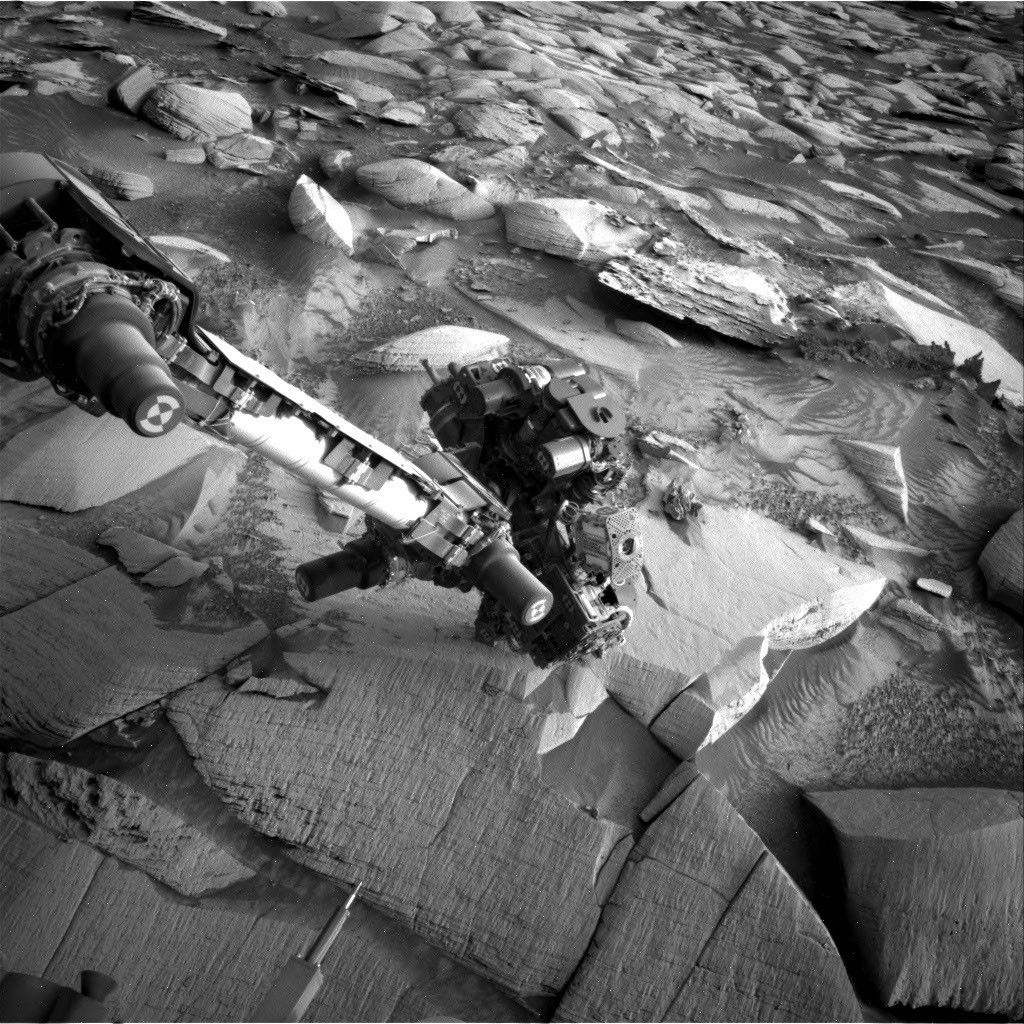 A black-and-white photograph taken by the Curiosity rover on Mars. The rover's mechanical arm extends from the left side of the frame toward the center, revealing a complex cluster of scientific instruments at its end. Below the arm, the Martian terrain consists of large, flat slabs of cracked, textured bedrock. The background is filled with scattered, lighter-colored rocks and patches of dark sand with small wind ripples.