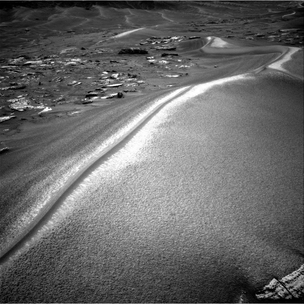 A black-and-white photograph from the Mars Curiosity rover showing a large, sweeping sand dune with a sharp, brightly sunlit crest. To the left are scattered flat rocks, and more rolling dunes stretch into the distant background.