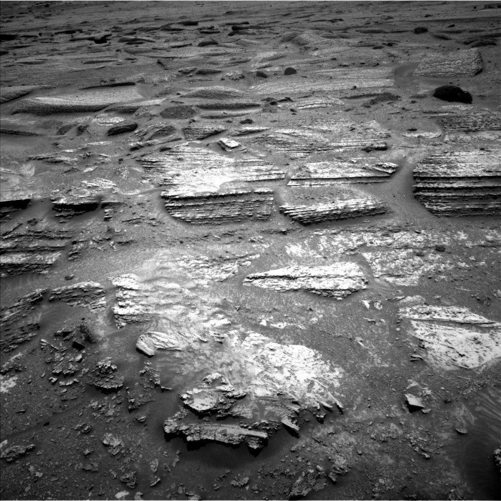 A black and white photograph of the Martian surface taken by the Curiosity rover, showing an expanse of fractured, light-colored bedrock. The rocks are distinctly layered and sedimentary, broken into flat, horizontal slabs and polygonal plates. Darker sand and loose dust fill the crevices and low areas between the rocky outcrops, emphasizing the rough, textured terrain.