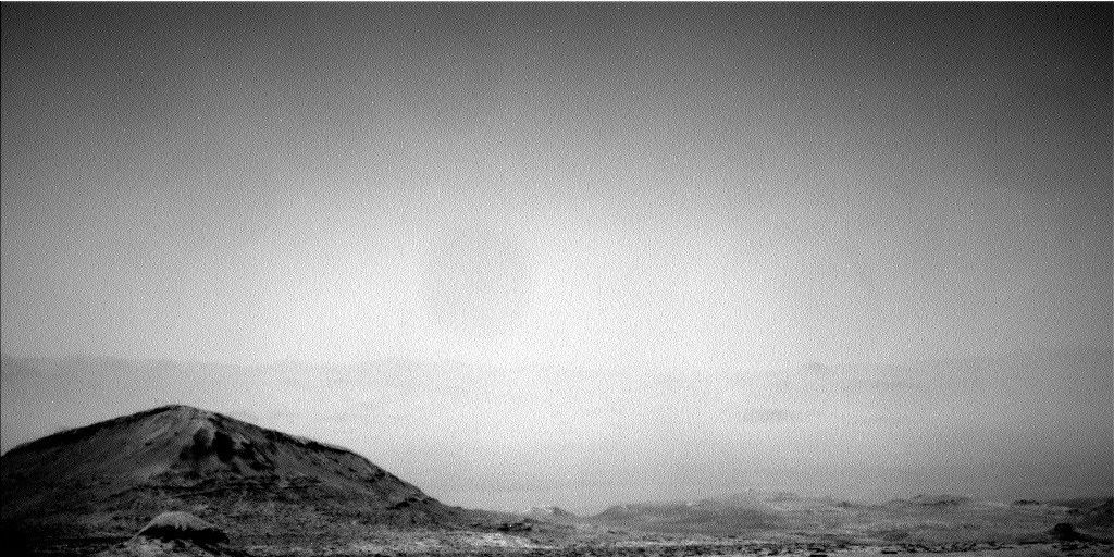 A grainy, black-and-white photograph of a barren Martian landscape taken by the Curiosity rover. In the lower left foreground, a prominent, dark hill with rugged textures and sloping sides rises above the terrain. The ground to the right is rocky and uneven, stretching into the distance to the faint, hazy outline of a jagged horizon line. The upper two-thirds of the image is filled with a bright, featureless, gray and hazy sky.