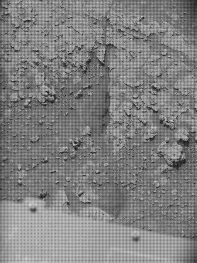 A grayscale photo of the surface of Mars shows rough, rocky terrain — knobby, jagged, and racked areas in the top half of the frame, with smoother areas dotted with numerous small rocks in the lower half. Part of the Curiosity rover is visible at the bottom of the frame.