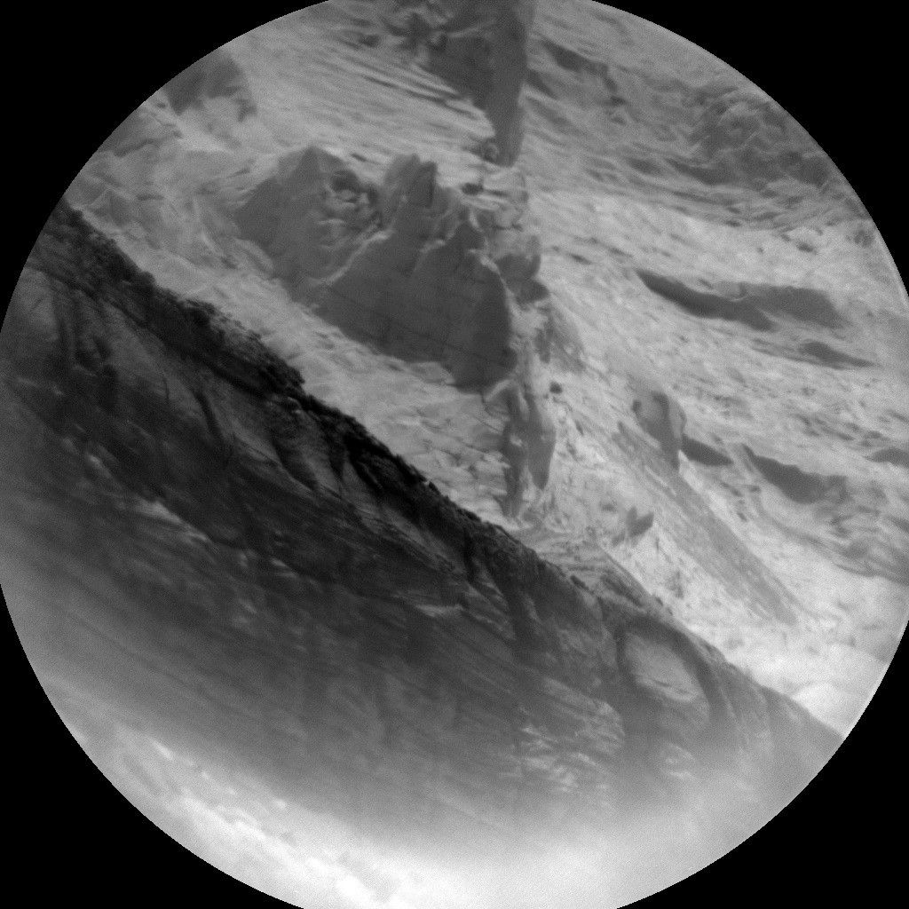 A round grayscale photograph from the Martian surface shows a close-up of jagged gray rocks. The slightly darker lower half of the frame, a band separated from the top of the image along a line extending from the upper left to the lower right of the image, features criss-crossing diagonal lines, and resembles a tire tread. The top half, lighter gray, features peaked, folded, and layered rocks that make that area appear like a tiny mountain range.