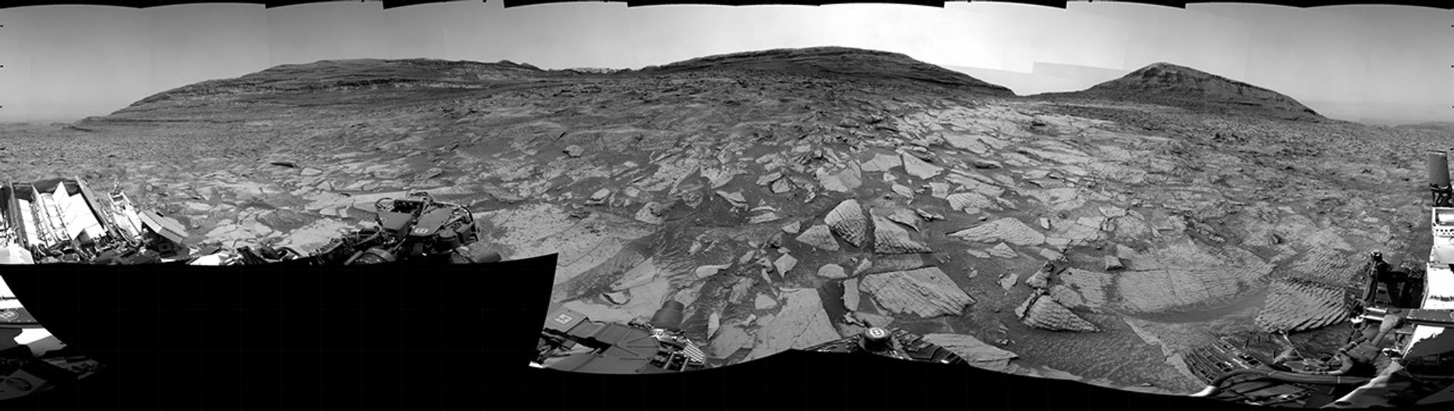 A grayscale panorama of the Martian surface, possibly at night or in twilight, shows a wide field of flat, dark gray terrain dotted with multiple jagged, medium-sized rocks stretching into the distance, where hills rise from the ground on the horizon. At center-right, a bright light behind a hill illuminates part of the sky, while the rest of the scene is fairly dark. Portions of the Curiosity rover are visible along the bottom of the image, including wheels in the lower-right corner.