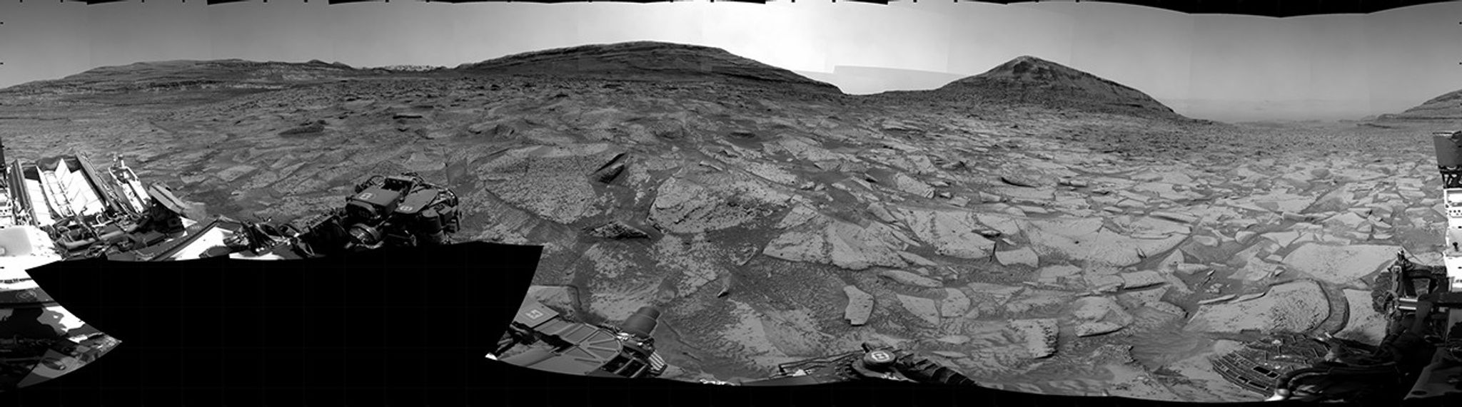 A grayscale panorama of the Martian surface, possibly at night or in twilight, shows a wide field of flat, dark gray terrain dotted with multiple jagged, medium-sized rocks stretching into the distance, where hills rise from the ground on the horizon. At center-right, a bright light behind a hill illuminates part of the sky, while the rest of the scene is fairly dark. Portions of the Curiosity rover are visible along the bottom of the image, including wheels in the lower-right corner.