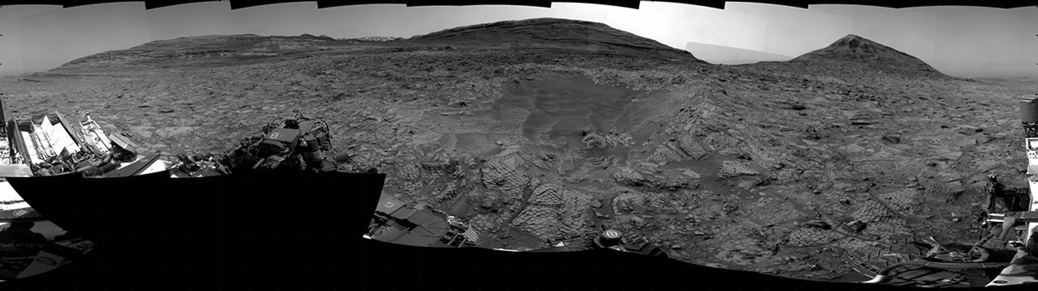 A grayscale panorama of the Martian surface, possibly at night or in twilight, shows a wide field of flat, dark gray terrain dotted with multiple jagged, medium-sized rocks stretching into the distance, where hills rise from the ground on the horizon. At center-right, a bright light behind a hill illuminates part of the sky, while the rest of the scene is fairly dark. Portions of the Curiosity rover are visible along the bottom of the image, including wheels in the lower-right corner.