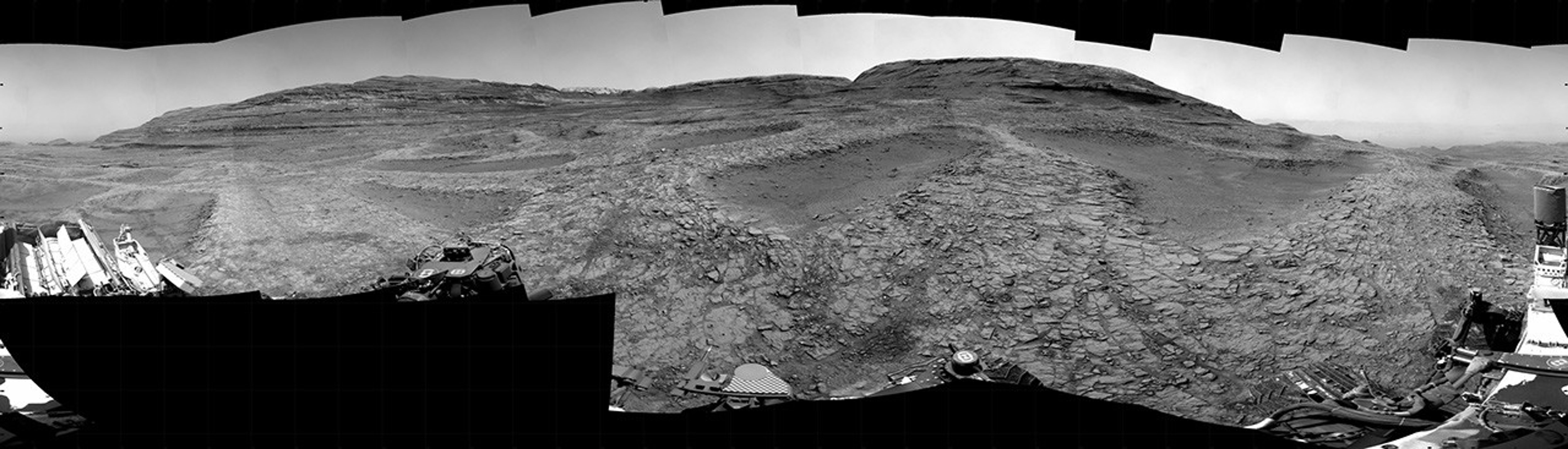 A grayscale panorama of the Martian surface, possibly at night or in twilight, shows a wide field of flat, dark gray terrain dotted with multiple jagged, medium-sized rocks stretching into the distance, where hills rise from the ground on the horizon. At center-right, a bright light behind a hill illuminates part of the sky, while the rest of the scene is fairly dark. Portions of the Curiosity rover are visible along the bottom of the image, including wheels in the lower-right corner.