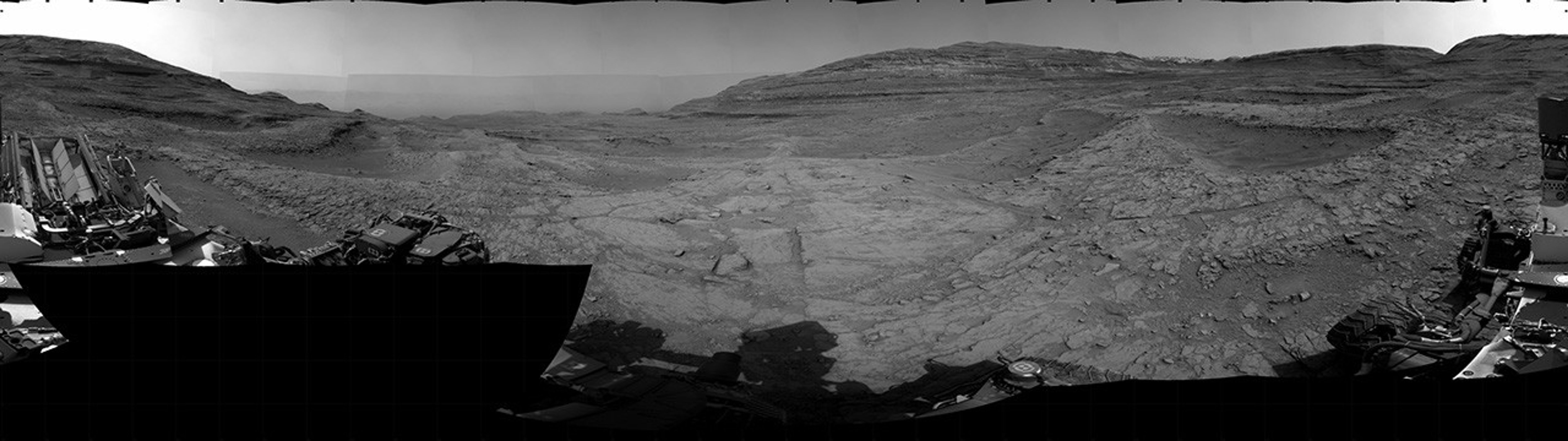 A grayscale panorama of the Martian surface, possibly at night or in twilight, shows a wide field of flat, dark gray terrain dotted with multiple jagged, medium-sized rocks stretching into the distance, where hills rise from the ground on the horizon. At center-right, a bright light behind a hill illuminates part of the sky, while the rest of the scene is fairly dark. Portions of the Curiosity rover are visible along the bottom of the image, including wheels in the lower-right corner.