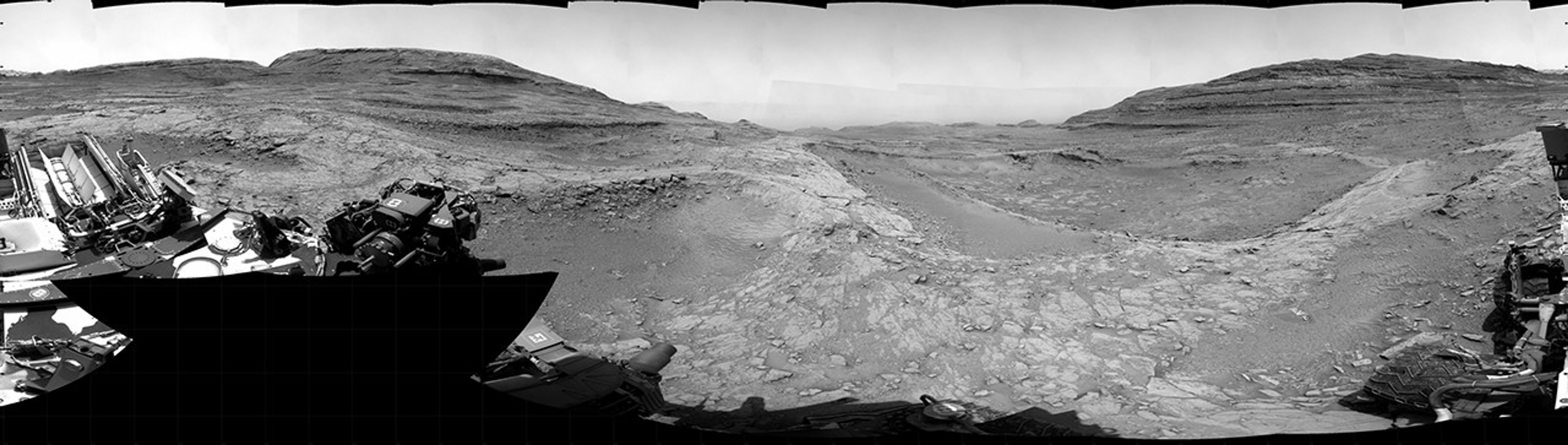 A grayscale panorama of the Martian surface, possibly at night or in twilight, shows a wide field of flat, dark gray terrain dotted with multiple jagged, medium-sized rocks stretching into the distance, where hills rise from the ground on the horizon. At center-right, a bright light behind a hill illuminates part of the sky, while the rest of the scene is fairly dark. Portions of the Curiosity rover are visible along the bottom of the image, including wheels in the lower-right corner.