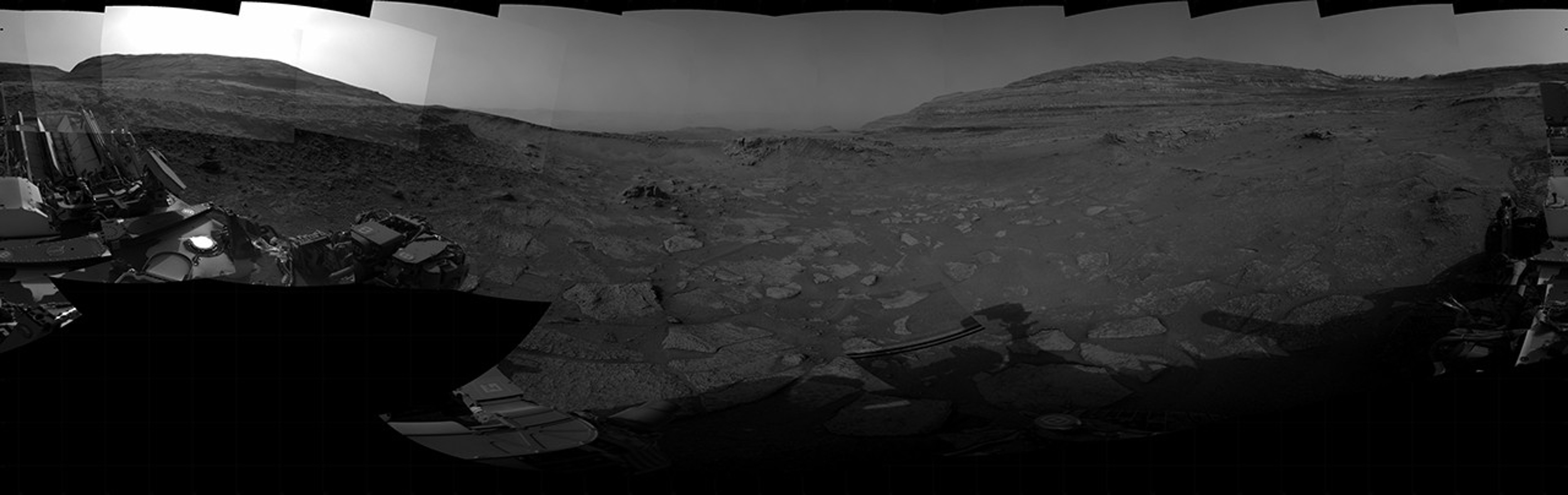 A grayscale panorama of the Martian surface, possibly at night or in twilight, shows a wide field of flat, dark gray terrain dotted with multiple jagged, medium-sized rocks stretching into the distance, where hills rise from the ground on the horizon. At center-right, a bright light behind a hill illuminates part of the sky, while the rest of the scene is fairly dark. Portions of the Curiosity rover are visible along the bottom of the image, including wheels in the lower-right corner.