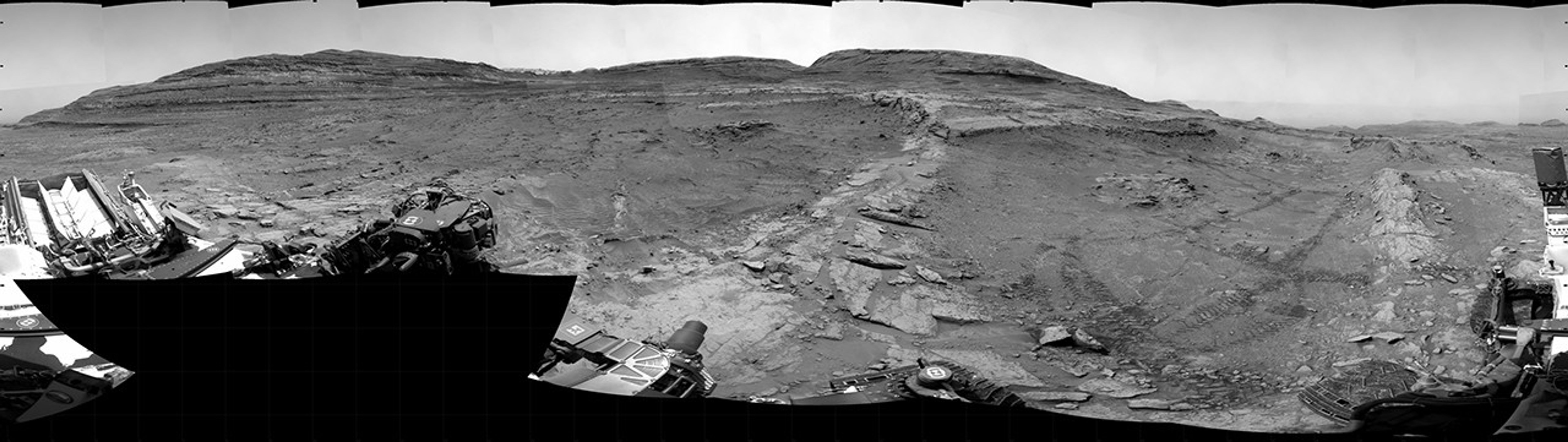 A grayscale panorama of the Martian surface, possibly at night or in twilight, shows a wide field of flat, dark gray terrain dotted with multiple jagged, medium-sized rocks stretching into the distance, where hills rise from the ground on the horizon. At center-right, a bright light behind a hill illuminates part of the sky, while the rest of the scene is fairly dark. Portions of the Curiosity rover are visible along the bottom of the image, including wheels in the lower-right corner.