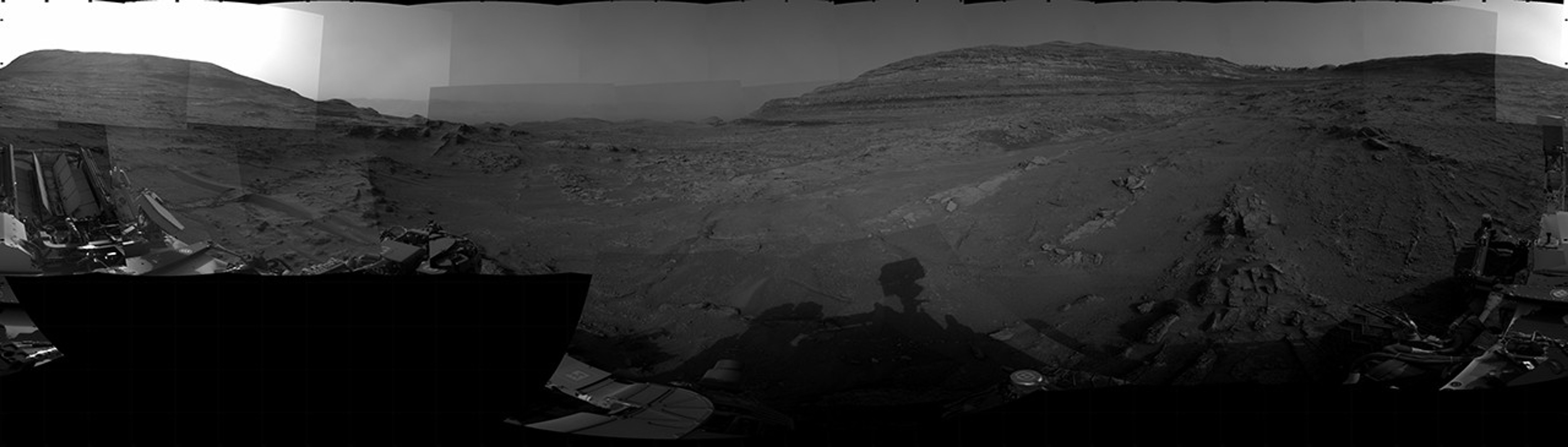 A grayscale panorama of the Martian surface, possibly at night or in twilight, shows a wide field of flat, dark gray terrain dotted with multiple jagged, medium-sized rocks stretching into the distance, where hills rise from the ground on the horizon. At center-right, a bright light behind a hill illuminates part of the sky, while the rest of the scene is fairly dark. Portions of the Curiosity rover are visible along the bottom of the image, including wheels in the lower-right corner.