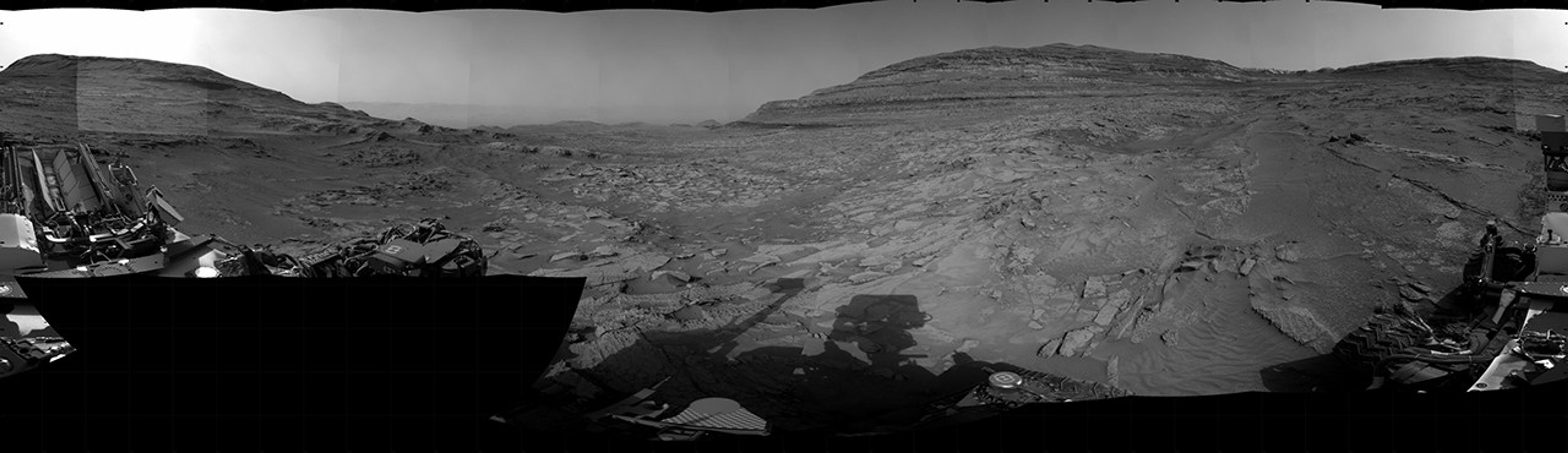 A grayscale panorama of the Martian surface, possibly at night or in twilight, shows a wide field of flat, dark gray terrain dotted with multiple jagged, medium-sized rocks stretching into the distance, where hills rise from the ground on the horizon. At center-right, a bright light behind a hill illuminates part of the sky, while the rest of the scene is fairly dark. Portions of the Curiosity rover are visible along the bottom of the image, including wheels in the lower-right corner.