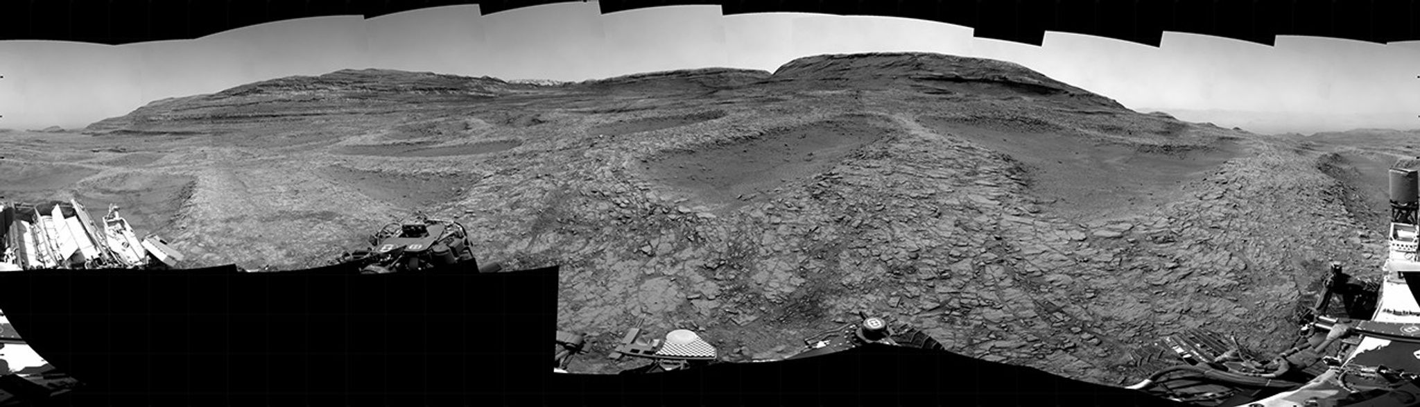 A grayscale panorama of the Martian surface, possibly at night or in twilight, shows a wide field of flat, dark gray terrain dotted with multiple jagged, medium-sized rocks stretching into the distance, where hills rise from the ground on the horizon. At center-right, a bright light behind a hill illuminates part of the sky, while the rest of the scene is fairly dark. Portions of the Curiosity rover are visible along the bottom of the image, including wheels in the lower-right corner.