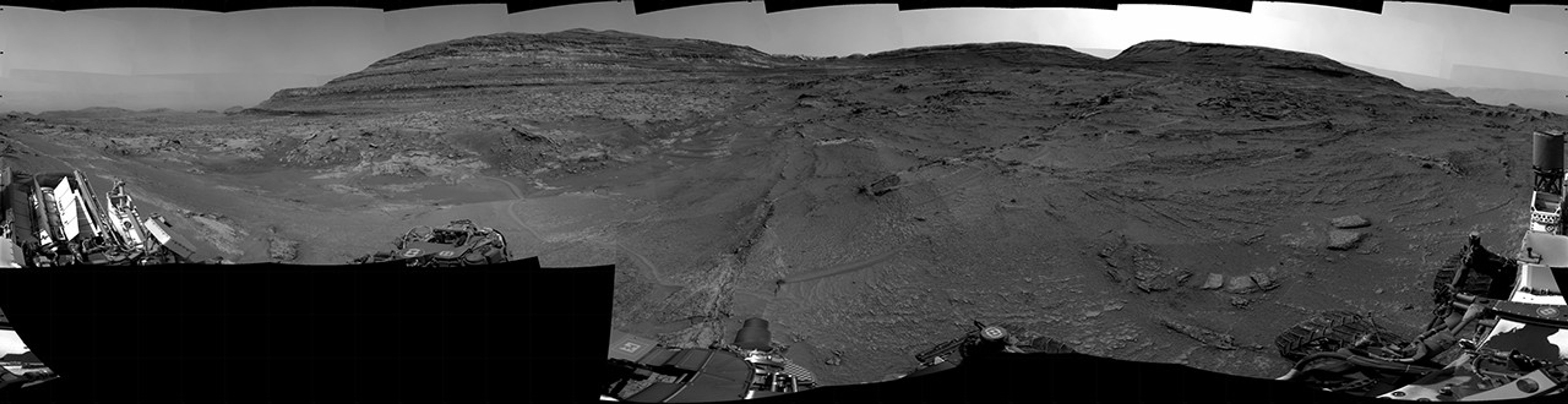 A grayscale panorama of the Martian surface, possibly at night or in twilight, shows a wide field of flat, dark gray terrain dotted with multiple jagged, medium-sized rocks stretching into the distance, where hills rise from the ground on the horizon. At center-right, a bright light behind a hill illuminates part of the sky, while the rest of the scene is fairly dark. Portions of the Curiosity rover are visible along the bottom of the image, including wheels in the lower-right corner.