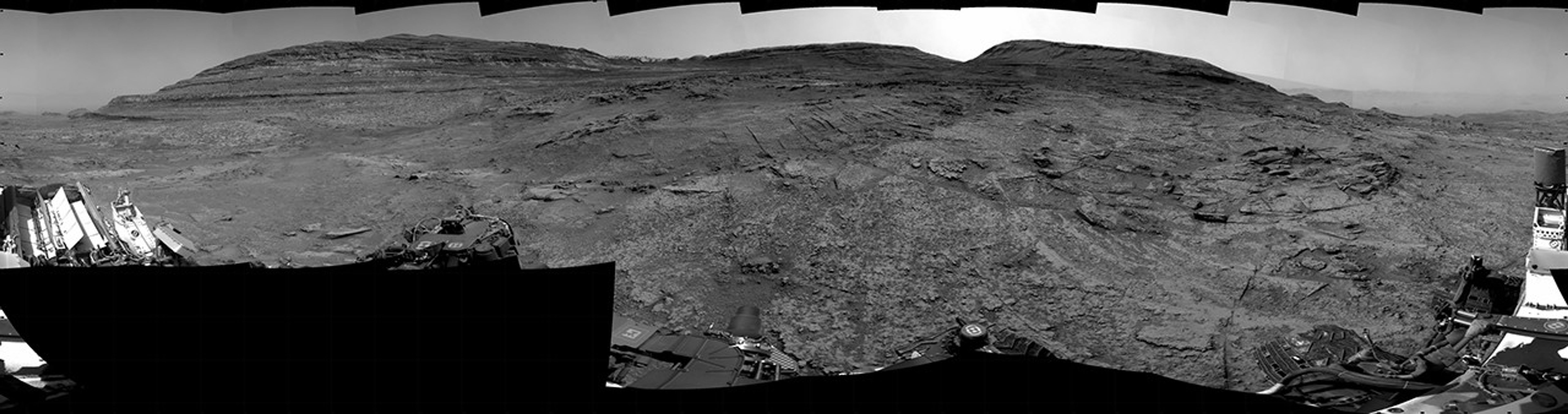 A grayscale panorama of the Martian surface, possibly at night or in twilight, shows a wide field of flat, dark gray terrain dotted with multiple jagged, medium-sized rocks stretching into the distance, where hills rise from the ground on the horizon. At center-right, a bright light behind a hill illuminates part of the sky, while the rest of the scene is fairly dark. Portions of the Curiosity rover are visible along the bottom of the image, including wheels in the lower-right corner.