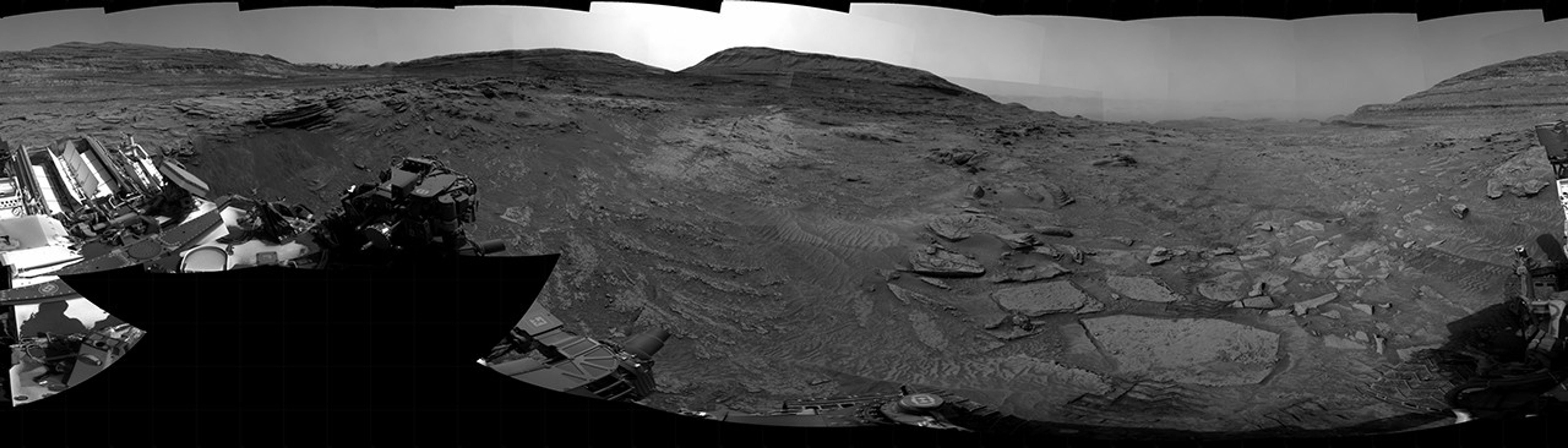 A grayscale panorama of the Martian surface, possibly at night or in twilight, shows a wide field of flat, dark gray terrain dotted with multiple jagged, medium-sized rocks stretching into the distance, where hills rise from the ground on the horizon. At center-right, a bright light behind a hill illuminates part of the sky, while the rest of the scene is fairly dark. Portions of the Curiosity rover are visible along the bottom of the image, including wheels in the lower-right corner.