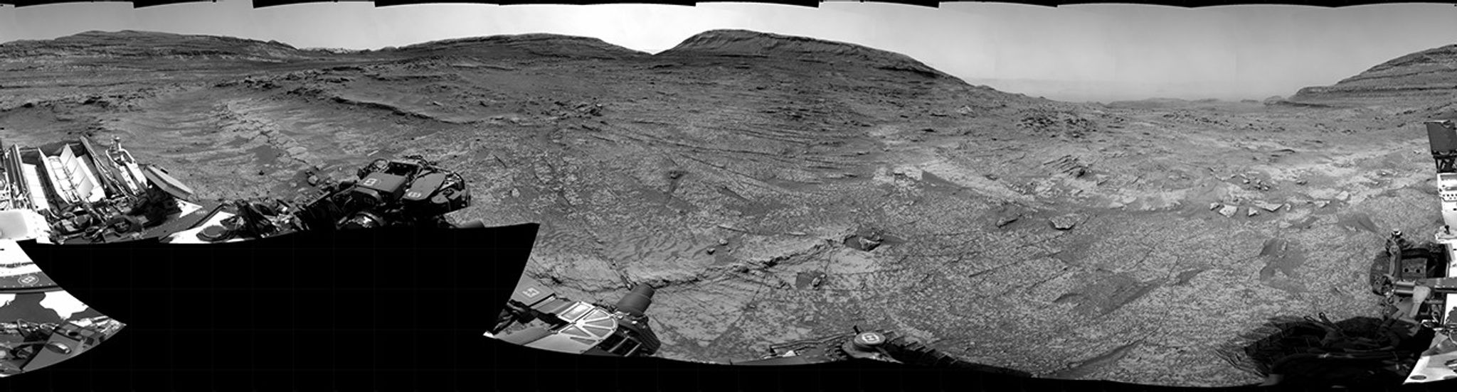 A grayscale panorama of the Martian surface, possibly at night or in twilight, shows a wide field of flat, dark gray terrain dotted with multiple jagged, medium-sized rocks stretching into the distance, where hills rise from the ground on the horizon. At center-right, a bright light behind a hill illuminates part of the sky, while the rest of the scene is fairly dark. Portions of the Curiosity rover are visible along the bottom of the image, including wheels in the lower-right corner.