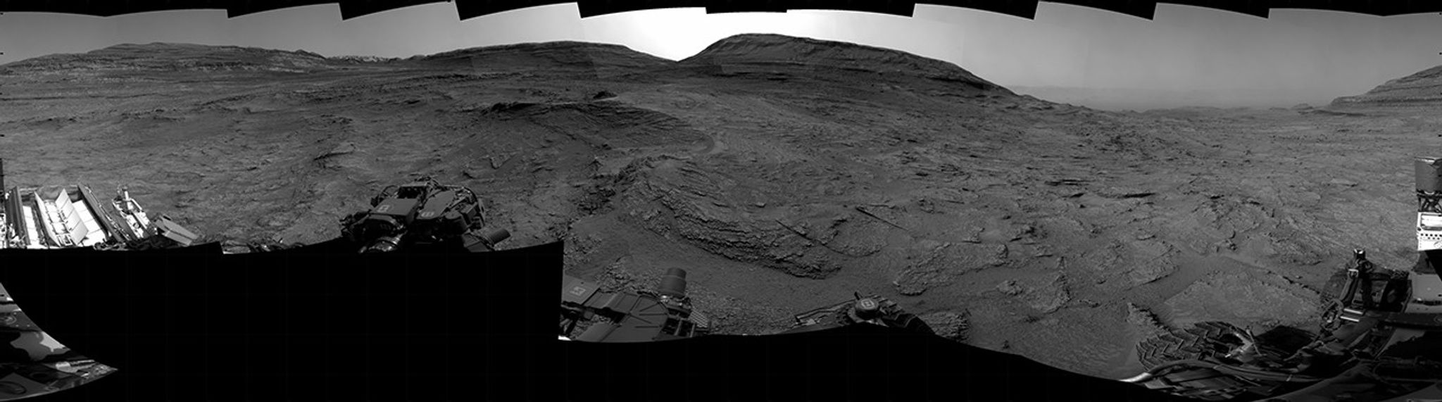 A grayscale panorama of the Martian surface, possibly at night or in twilight, shows a wide field of flat, dark gray terrain dotted with multiple jagged, medium-sized rocks stretching into the distance, where hills rise from the ground on the horizon. At center-right, a bright light behind a hill illuminates part of the sky, while the rest of the scene is fairly dark. Portions of the Curiosity rover are visible along the bottom of the image, including wheels in the lower-right corner.