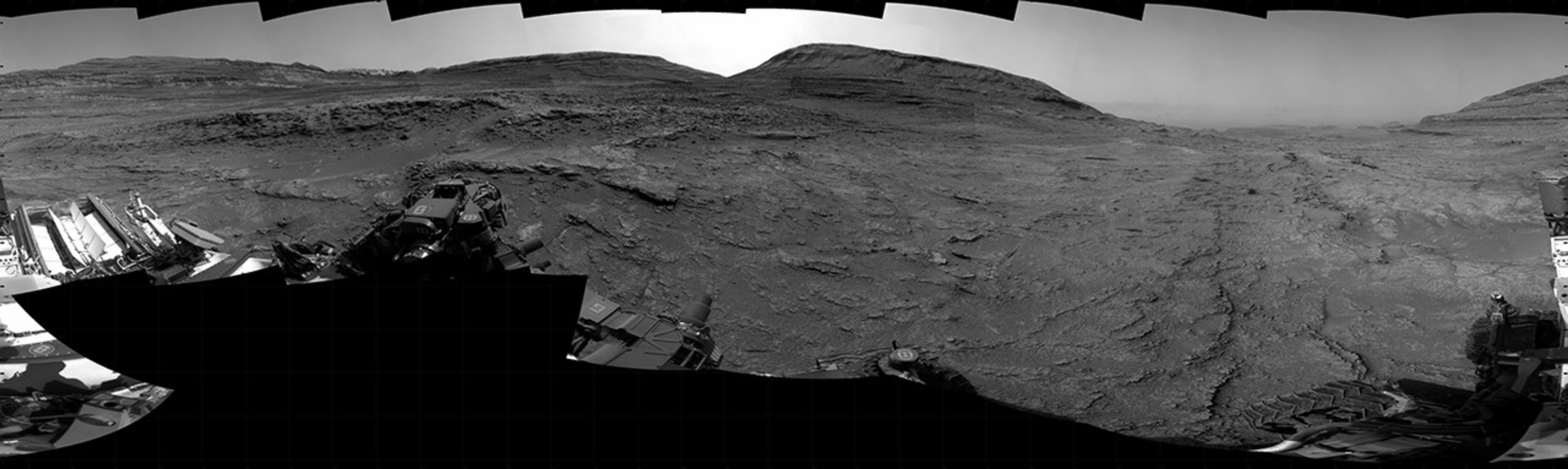 A grayscale panorama of the Martian surface, possibly at night or in twilight, shows a wide field of flat, dark gray terrain dotted with multiple jagged, medium-sized rocks stretching into the distance, where hills rise from the ground on the horizon. At center-right, a bright light behind a hill illuminates part of the sky, while the rest of the scene is fairly dark. Portions of the Curiosity rover are visible along the bottom of the image, including wheels in the lower-right corner.