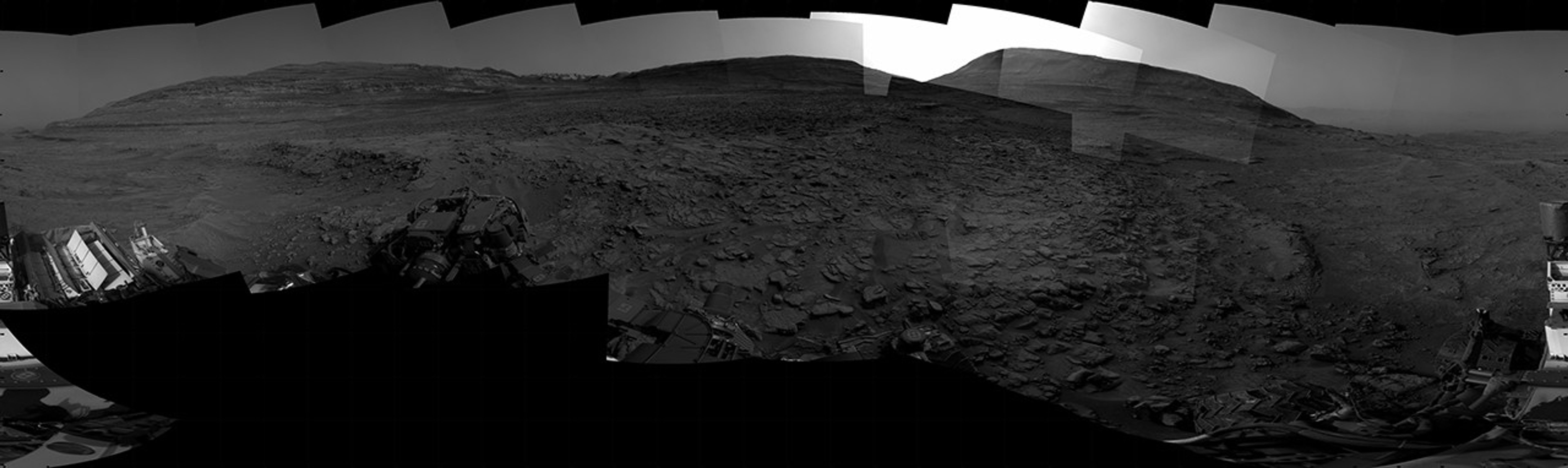 A grayscale panorama of the Martian surface, possibly at night or in twilight, shows a wide field of flat, dark gray terrain dotted with multiple jagged, medium-sized rocks stretching into the distance, where hills rise from the ground on the horizon. At center-right, a bright light behind a hill illuminates part of the sky, while the rest of the scene is fairly dark. Portions of the Curiosity rover are visible along the bottom of the image, including wheels in the lower-right corner.