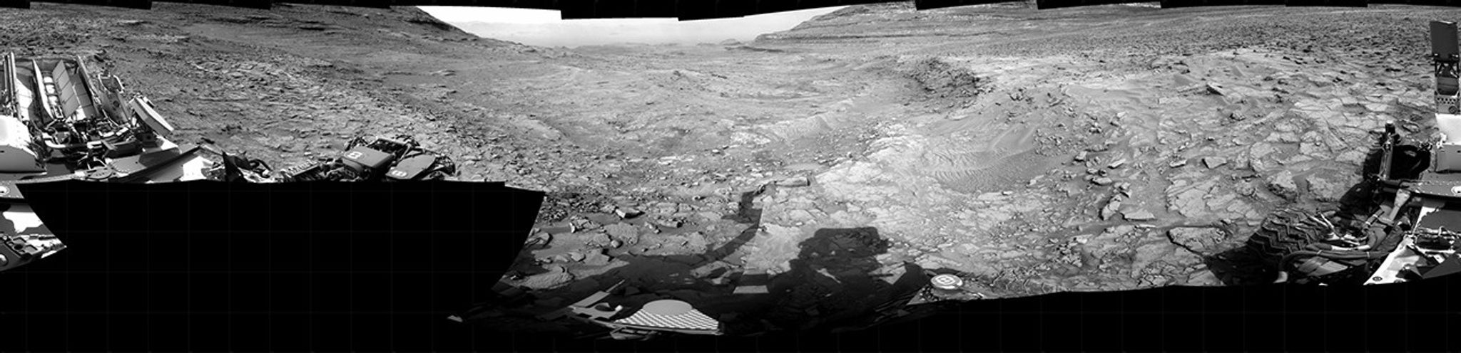 A grayscale panorama of the Martian surface, possibly at night or in twilight, shows a wide field of flat, dark gray terrain dotted with multiple jagged, medium-sized rocks stretching into the distance, where hills rise from the ground on the horizon. At center-right, a bright light behind a hill illuminates part of the sky, while the rest of the scene is fairly dark. Portions of the Curiosity rover are visible along the bottom of the image, including wheels in the lower-right corner.