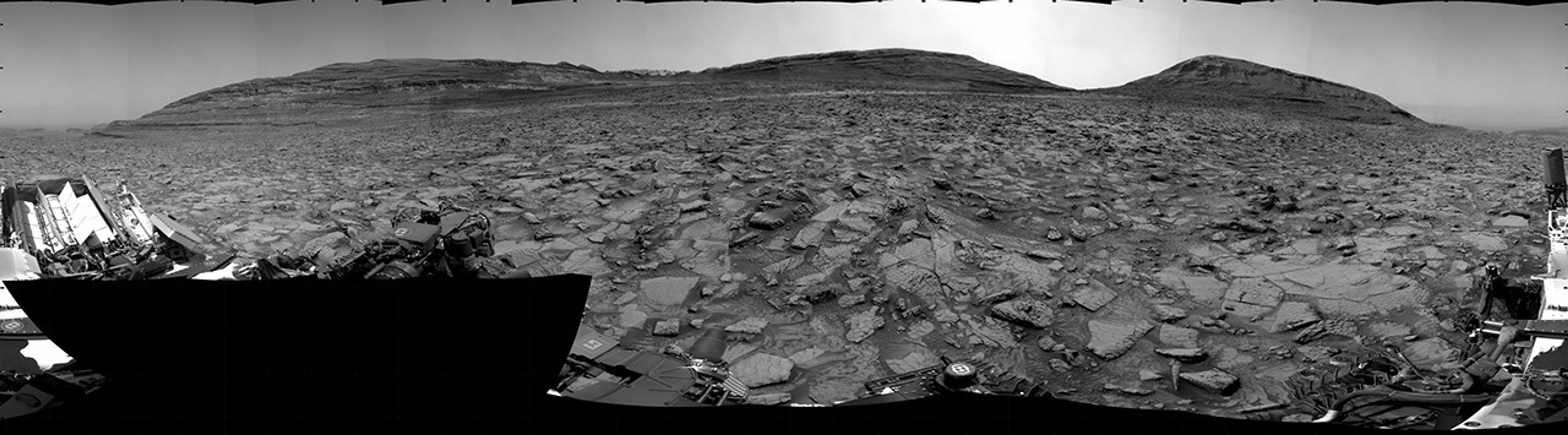 A grayscale panorama of the Martian surface, possibly at night or in twilight, shows a wide field of flat, dark gray terrain dotted with multiple jagged, medium-sized rocks stretching into the distance, where hills rise from the ground on the horizon. At center-right, a bright light behind a hill illuminates part of the sky, while the rest of the scene is fairly dark. Portions of the Curiosity rover are visible along the bottom of the image, including wheels in the lower-right corner.