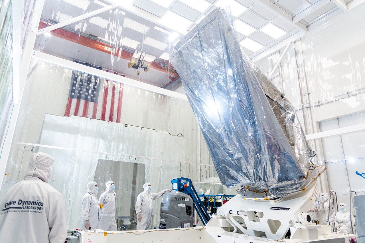 Inside a large white clean room, a large enclosure – part of the NEO Surveyor space telescope – sits at the center of the image, atop a platform or hydraulic lift on the floor that is holding it aloft at a 45-degree angle. The enclosure is wrapped inside what looks like a large clear plastic bag. A large American flag hangs on a wall in the background. at right. Workers dressed head-to-toe in white “bunny suit” protective clothing attend to the spacecraft enclosure, with one in the foreground, wearing glasses, looking up at it. A group of others is chatting in the background, beneath the flag. One other is in the background at far right, on the opposite end of the lift.