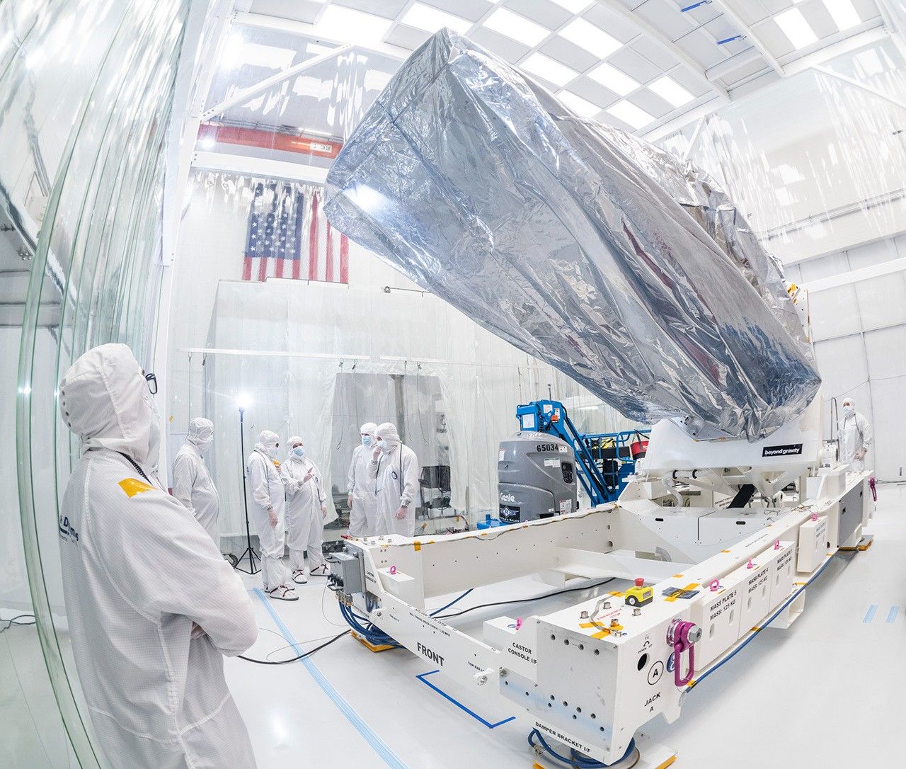 Inside a large white clean room, a large enclosure – part of the NEO Surveyor space telescope – sits at the center of the image, atop a platform or hydraulic lift on the floor that is holding it aloft at a 45-degree angle. The enclosure is wrapped inside what looks like a large clear plastic bag. A large American flag hangs on a wall in the background. at right. Workers dressed head-to-toe in white “bunny suit” protective clothing attend to the spacecraft enclosure, with one in the foreground, wearing glasses, looking up at it. A group of others is chatting in the background, beneath the flag. One other is in the background at far right, on the opposite end of the lift.