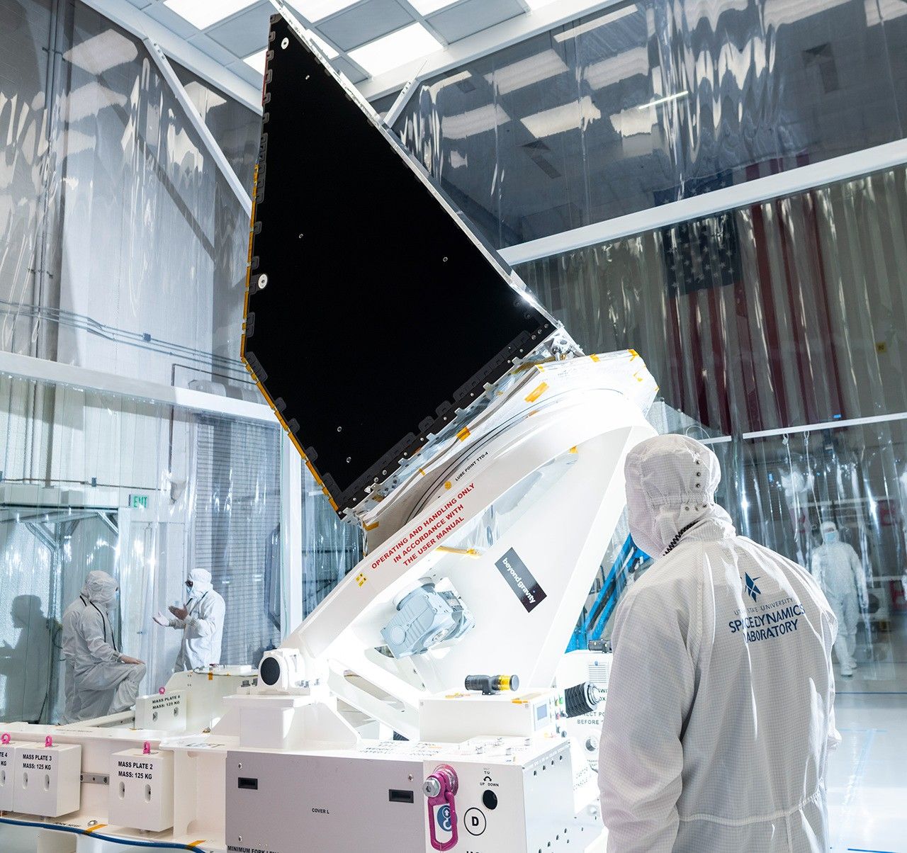 Inside a large white clean room, a large enclosure – part of the NEO Surveyor space telescope – sits at the center of the image, upright atop a platform on the floor. A large, shiny black panel faces left. A large American flag hangs on a wall at right. Workers dressed head-to-toe in white “bunny suit” protective clothing attend to the spacecraft asembly, with two chatting off to the left side, and one in the right foreground of the image looking at the enclosure from a distance. One other person is in the background, beneath the flag.