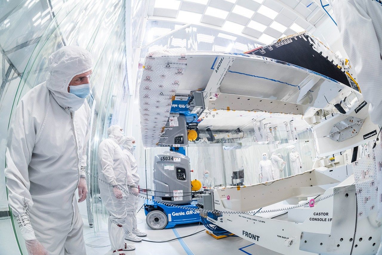 Inside a large white clean room, technicians are examining a large enclosure – part of the NEO Surveyor space telescope – at the center-right of the image, elevated atop a platform on the floor, so the bottom of the enclosure is above their heads. Three workers dressed head-to-toe in white “bunny suit” protective clothing attend to the spacecraft, eyeing it from the left side of the image. Others sit or stand in the background, behind the enclosure.