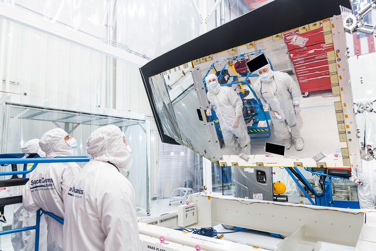 Inside a large white clean room, technicians are examining a large enclosure – part of the NEO Surveyor space telescope – at the center-right of the image, elevated atop a platform on the floor. Two workers dressed head-to-toe in white “bunny suit” protective clothing attend to the spacecraft; they are reflected in a mirror-like panel on the spacecraft assembly at right.