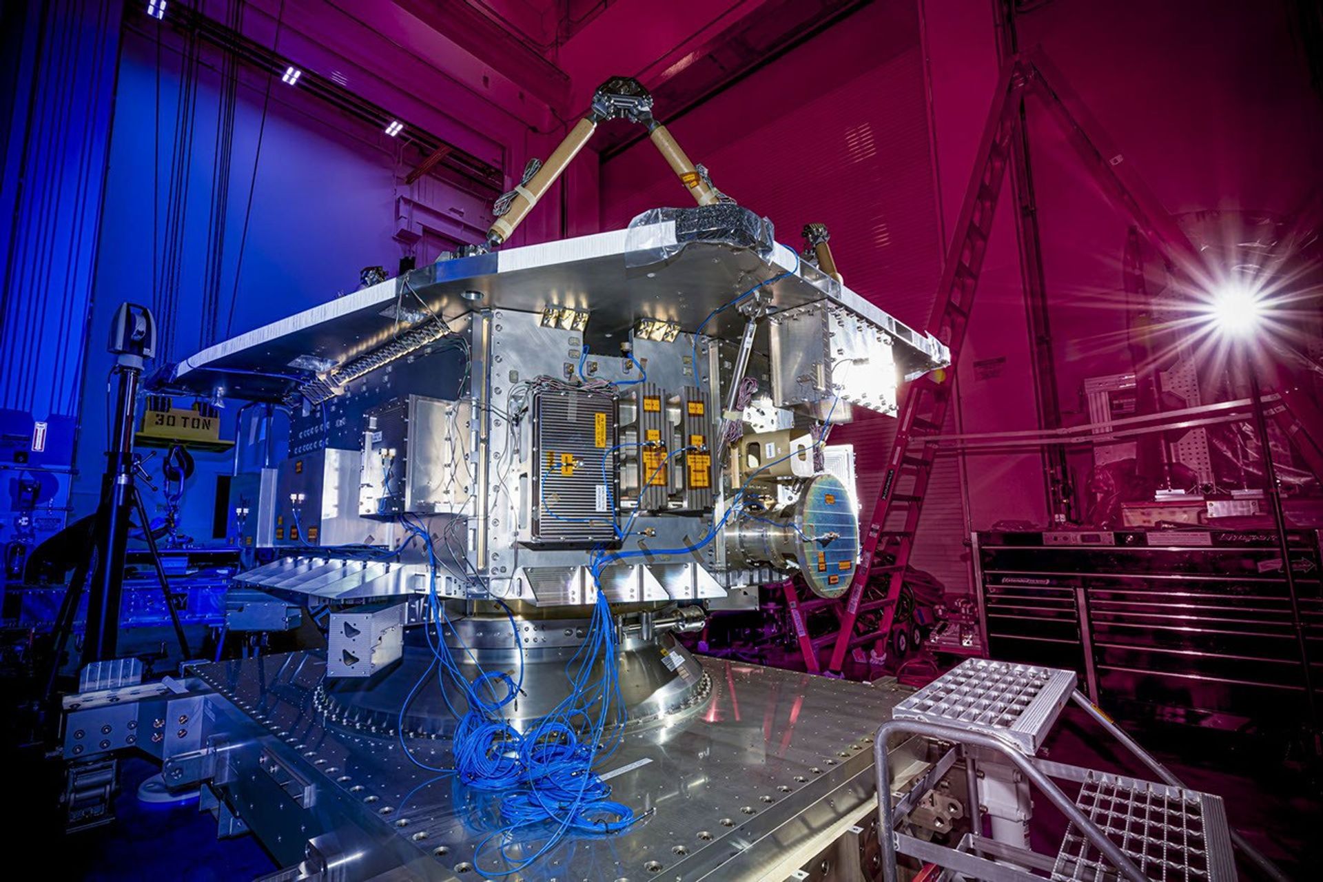 The bus structure of NASA’s NEO Surveyor sits on a platform during testing at BAE Systems, featuring a complex metallic assembly of power, propulsion, and communication subsystems under dramatic pink and blue lighting.