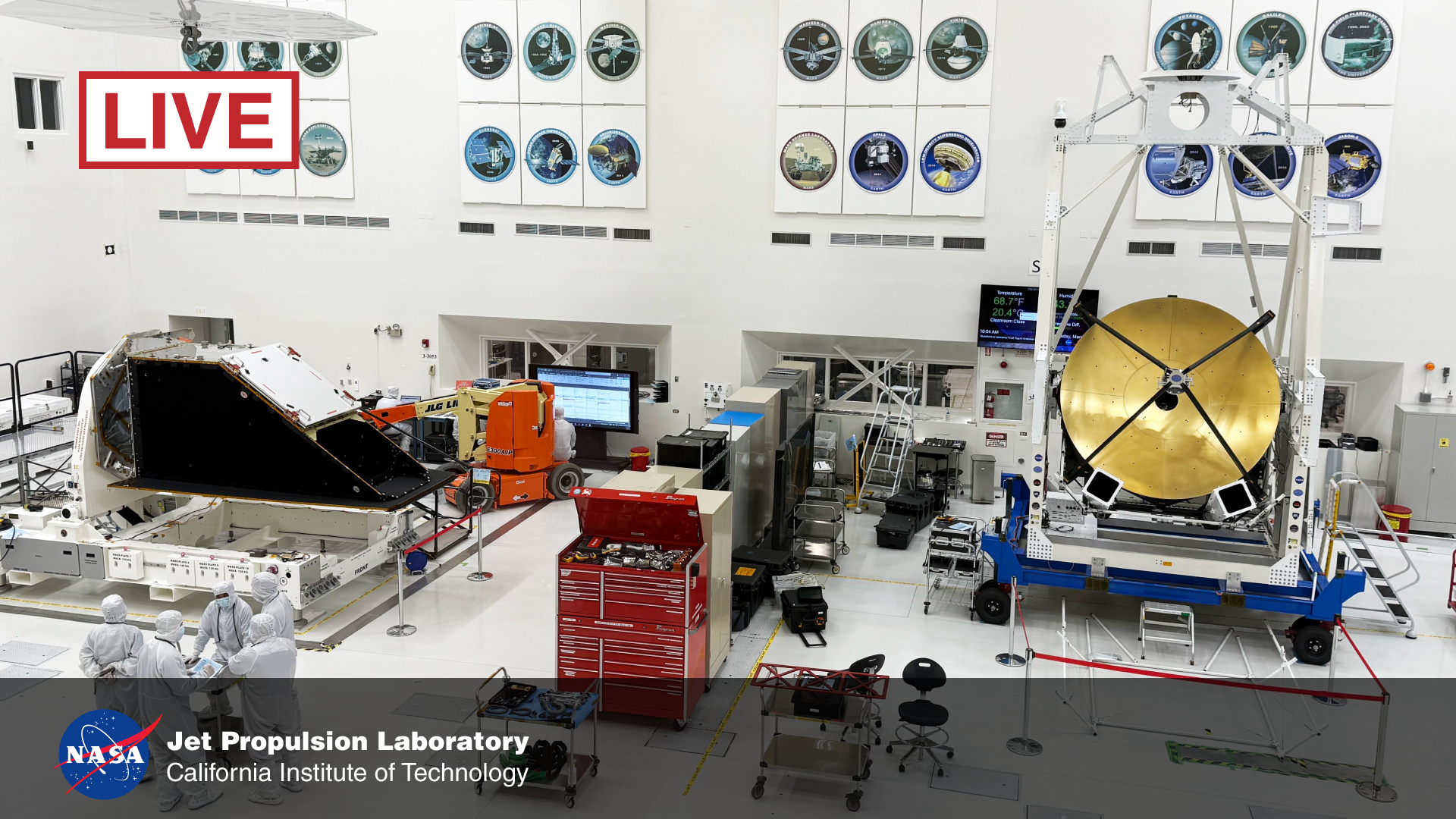 A color photo of a very large, white clean room, where two spacecraft are being assembled on the floor. A group of workers dressed head-to-toe in white sanitary suits is huddled in the foreground at left. Behind them is a large black enclosure, trapezoid-shaped, resting on its long side, with its angled side pointing to the right. On the angled face, the top half is covered in a bright, silvery white panel, while the bottom half is opened to the inside. On the far right of the room, the other spacecraft is inside a large white scaffold with a ring at its top, sitting on a car-sized blue cart with black wheels. About the only part of the craft visible is a very large gold dish, facing the viewer, with thin black supports criss-crossed in an X outside its opening, holding a small metallic box in the middle. On the floor between the two craft are various rolling chairs and carts, including a large red tool chest. High on the wall behind everything are arrayed 24 circles showing the insignias of previous spacecraft built in this room.