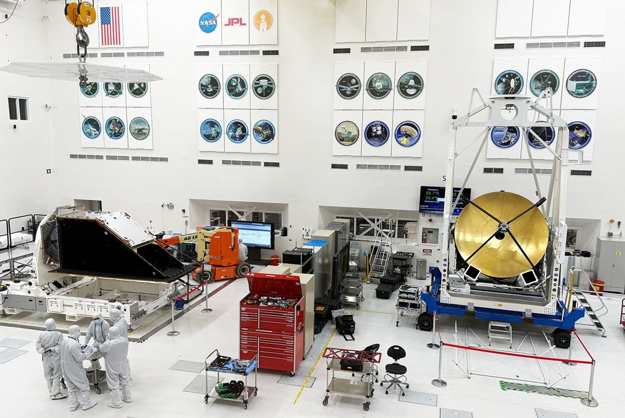 Workers in white protective clothing work on spacecraft in a large white room.