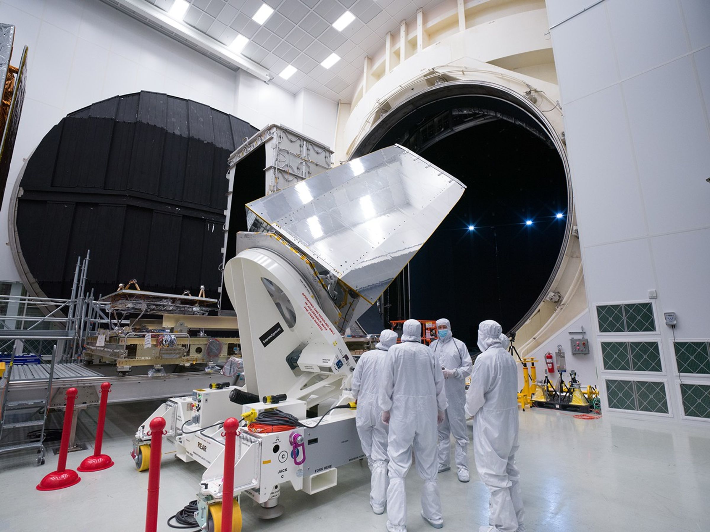 Workers in white protective clothing stand to the side of a white dolly holding NEO Surveyor's instrument enclosure. The side of the enclosure that is visible in this image is shiny, and metallic like.