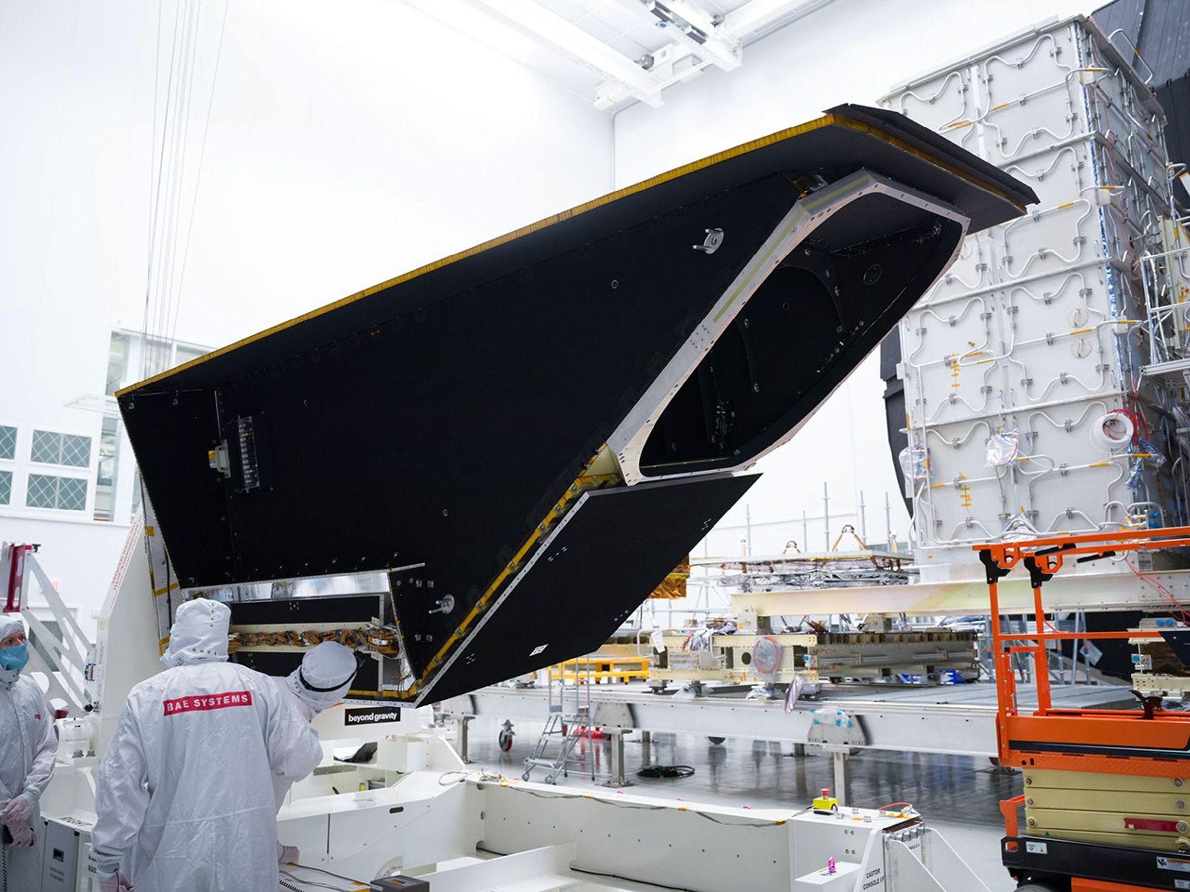 A large, black triangular-shaped object sits on a white platform with workers in white protective clothing nearby. An orange lift is to the right and lots of other equipment is in the background.