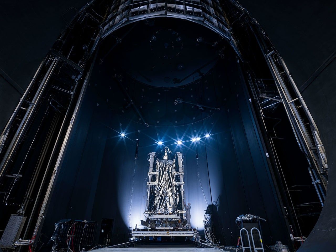 A piece of a spacecraft sits on a dolly with lights over it.