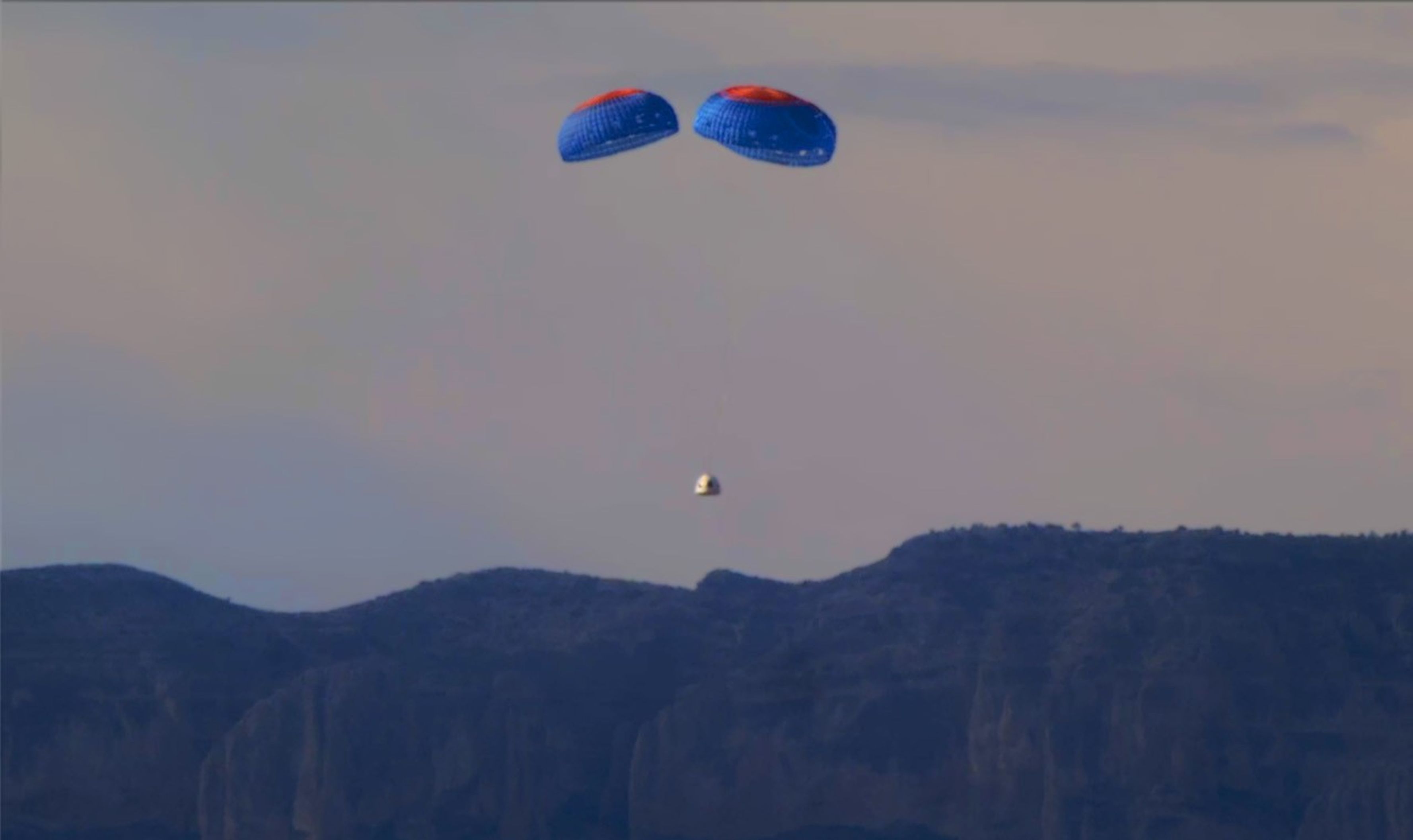 Small spacecraft suspended from three blue-and-red parachutes descends over a mountain ridgeline.