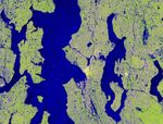 A false-color satellite image shows the Seattle metropolitan area, with deep blue water bodies like Puget Sound and Lake Washington contrasting against a textured, light green and purple urban landscape divided by thin transportation lines.