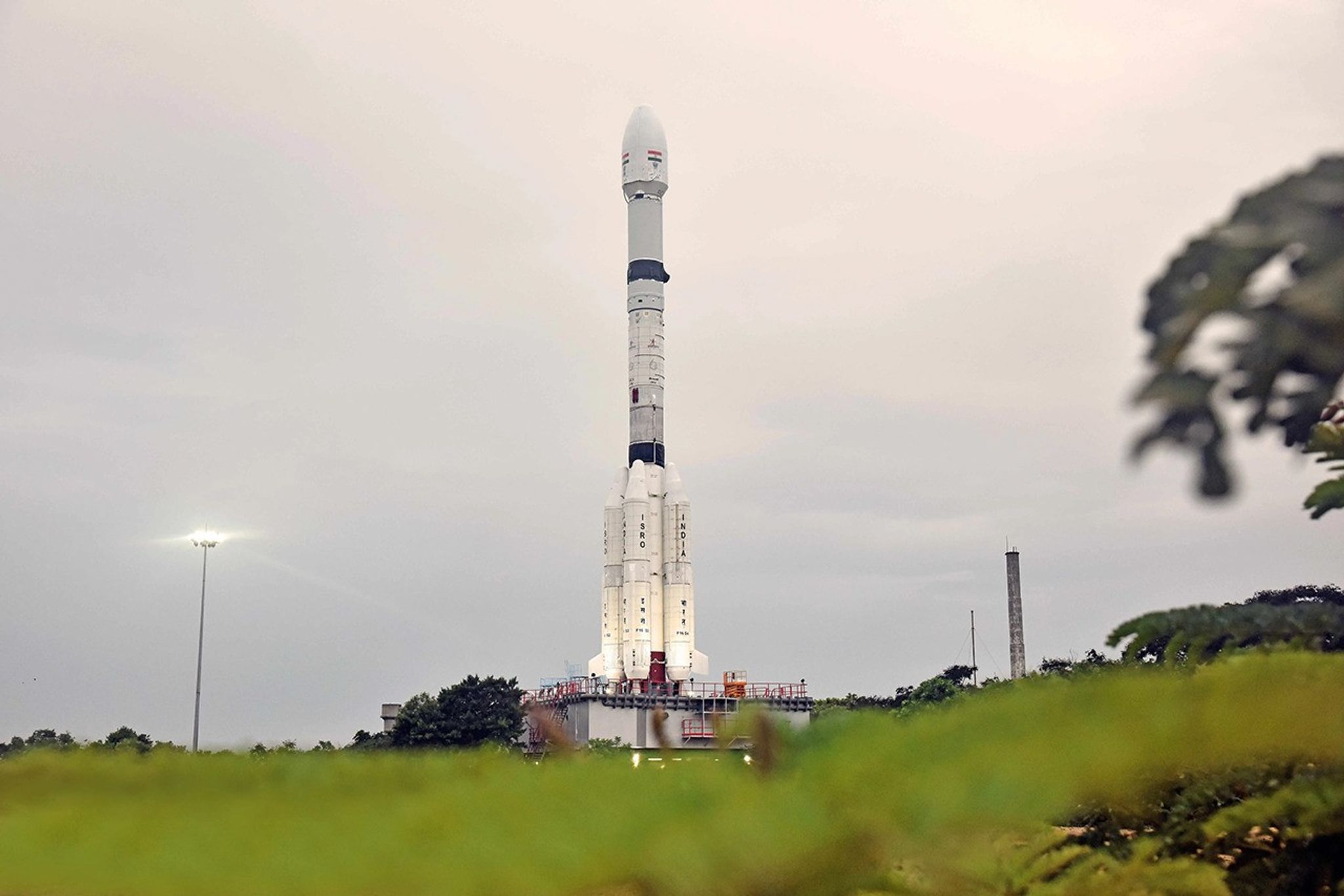 A low-angle, eye-level shot of the white NISAR satellite rocket on the launchpad. The rocket is tall and thin with "ISRO" and "INDIA" printed in red on its side, and the Indian flag is visible near the top. It stands against a cloudy, overcast sky, with lush green foliage out of focus in the foreground.