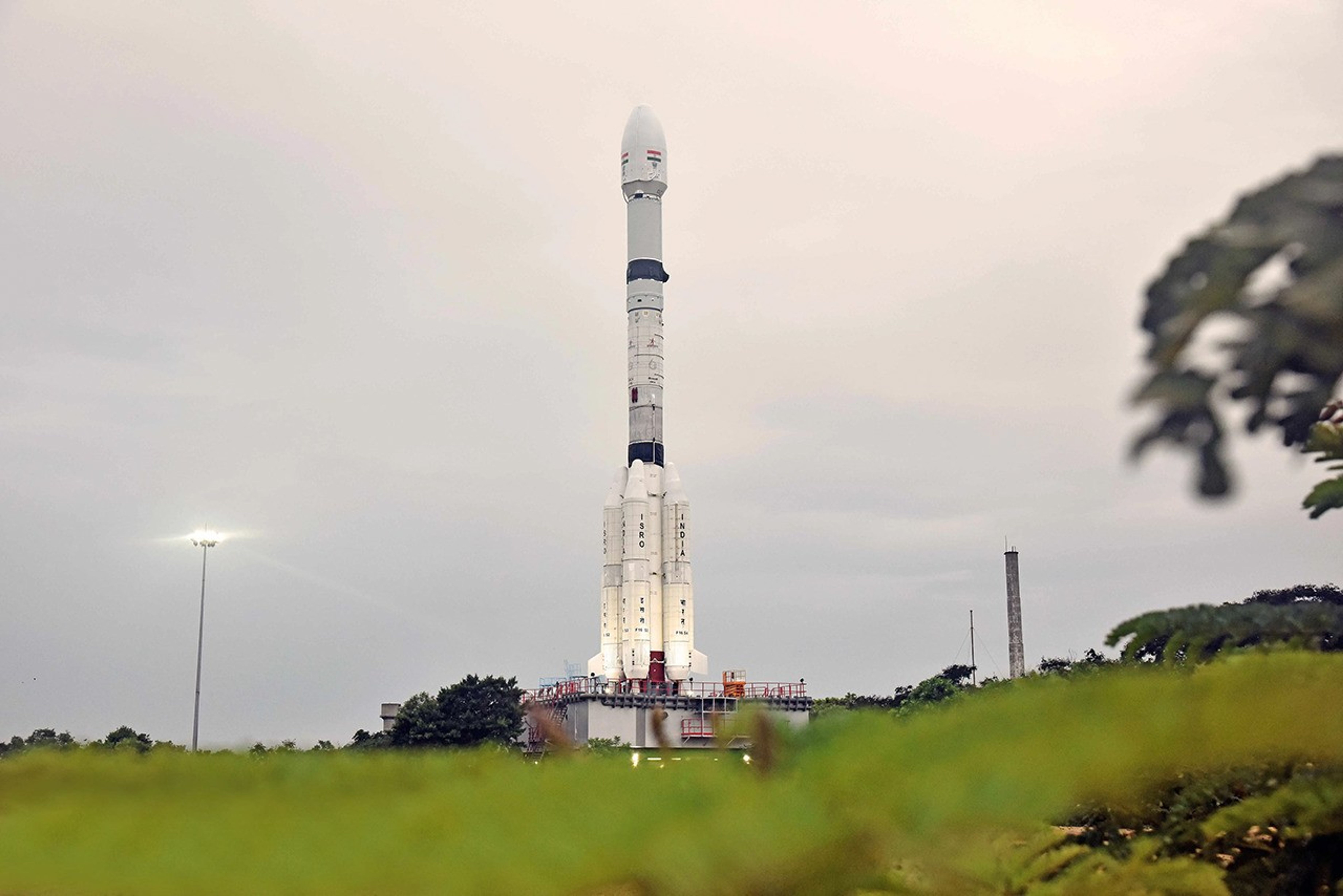 A low-angle, eye-level shot of the white NISAR satellite rocket on the launchpad. The rocket is tall and thin with "ISRO" and "INDIA" printed in red on its side, and the Indian flag is visible near the top. It stands against a cloudy, overcast sky, with lush green foliage out of focus in the foreground.