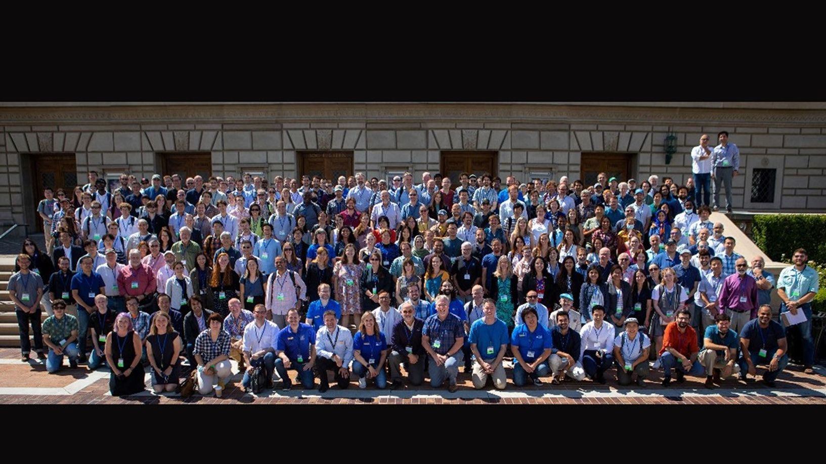 Large group photo of over 100 members of the NISAR mission team gathered outdoors on steps in front of a stone building, smiling for the camera.